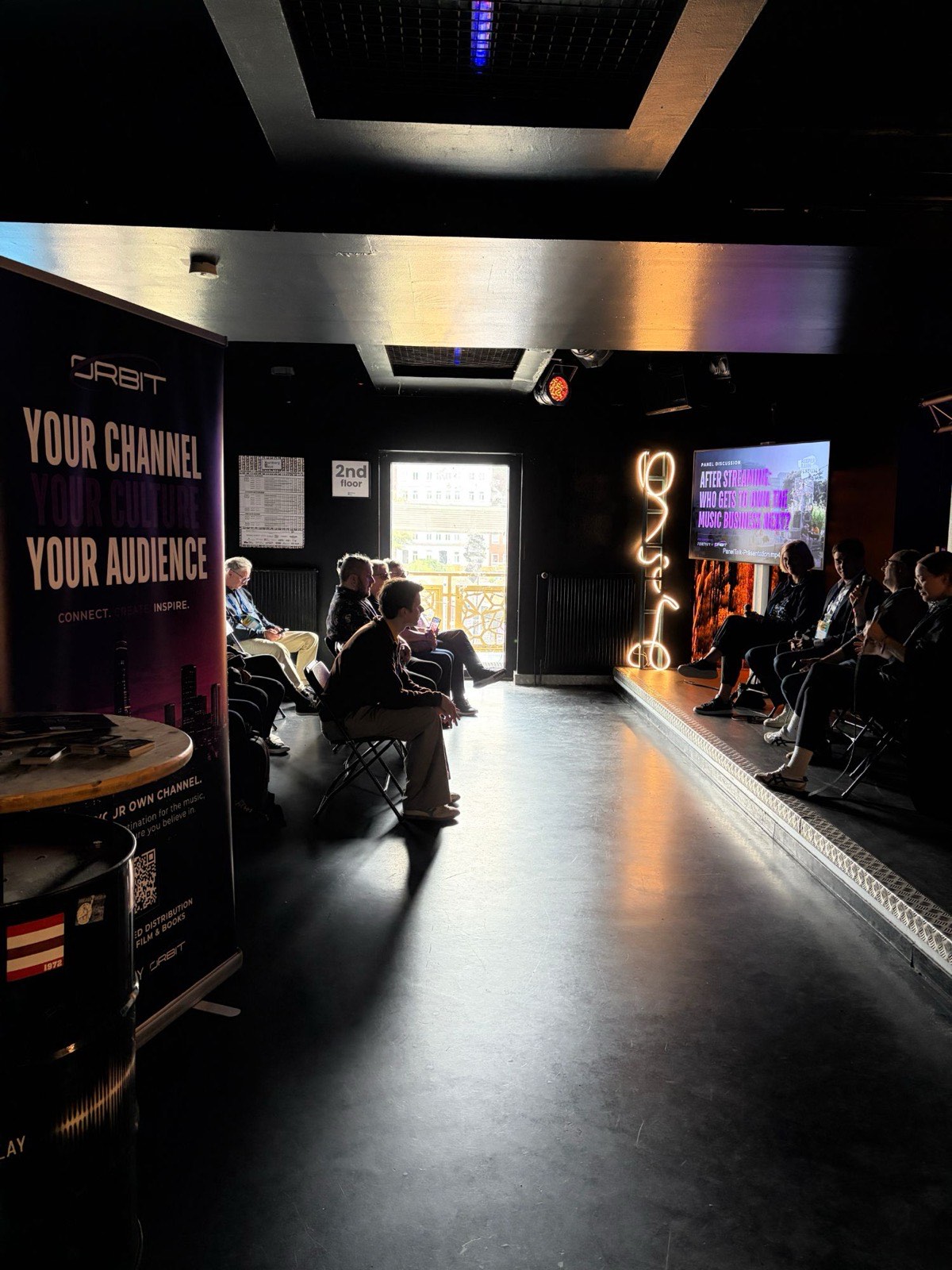 The Twentyfive Club room before the panel — an Orbit banner reading “Your channel, your culture, your audience” on the left, the seated audience in the middle, and the lit stage on the right