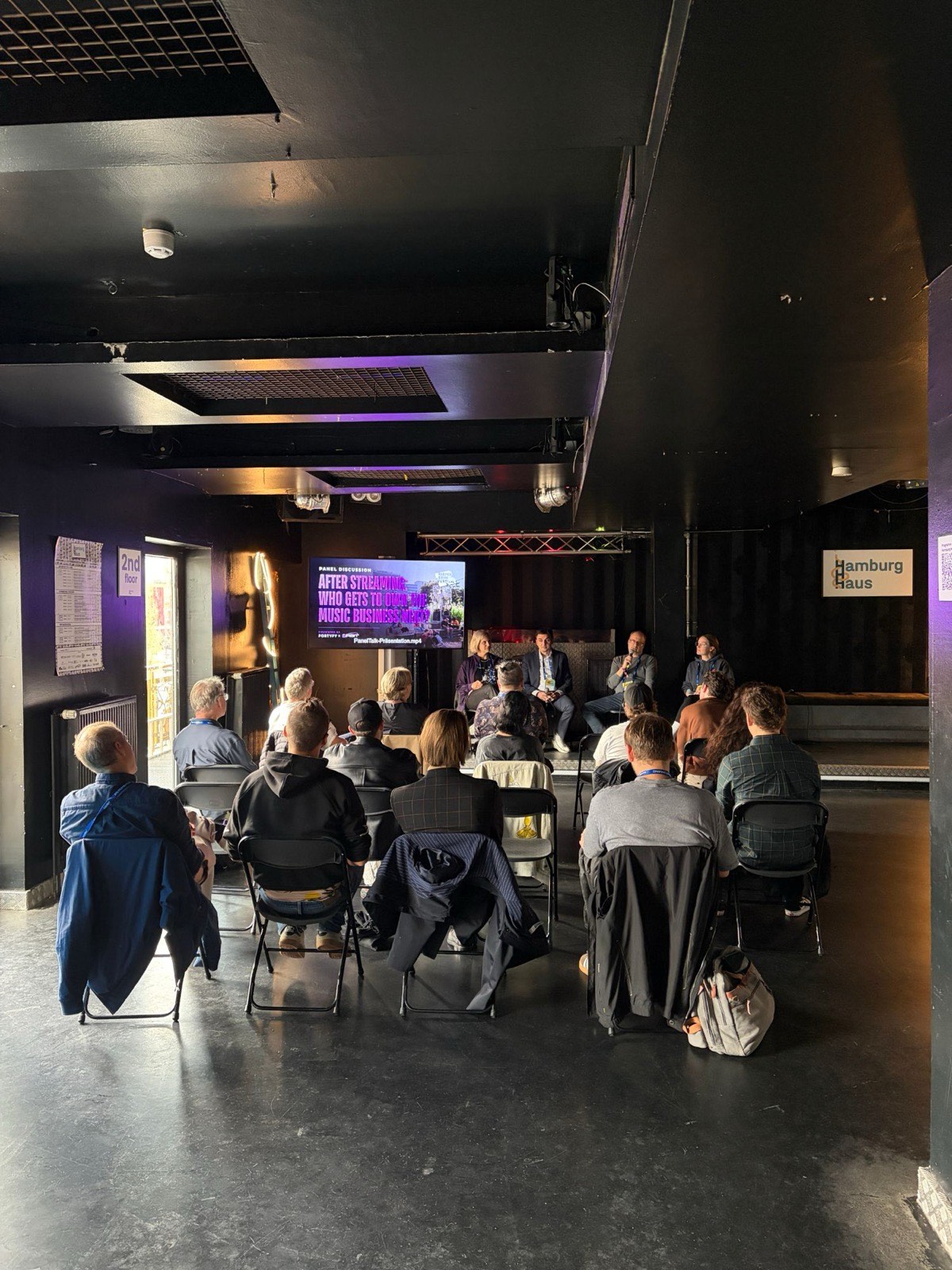 The full room seen from the back: rows of folding chairs facing the four panellists on the low stage, the title slide on the screen beside them