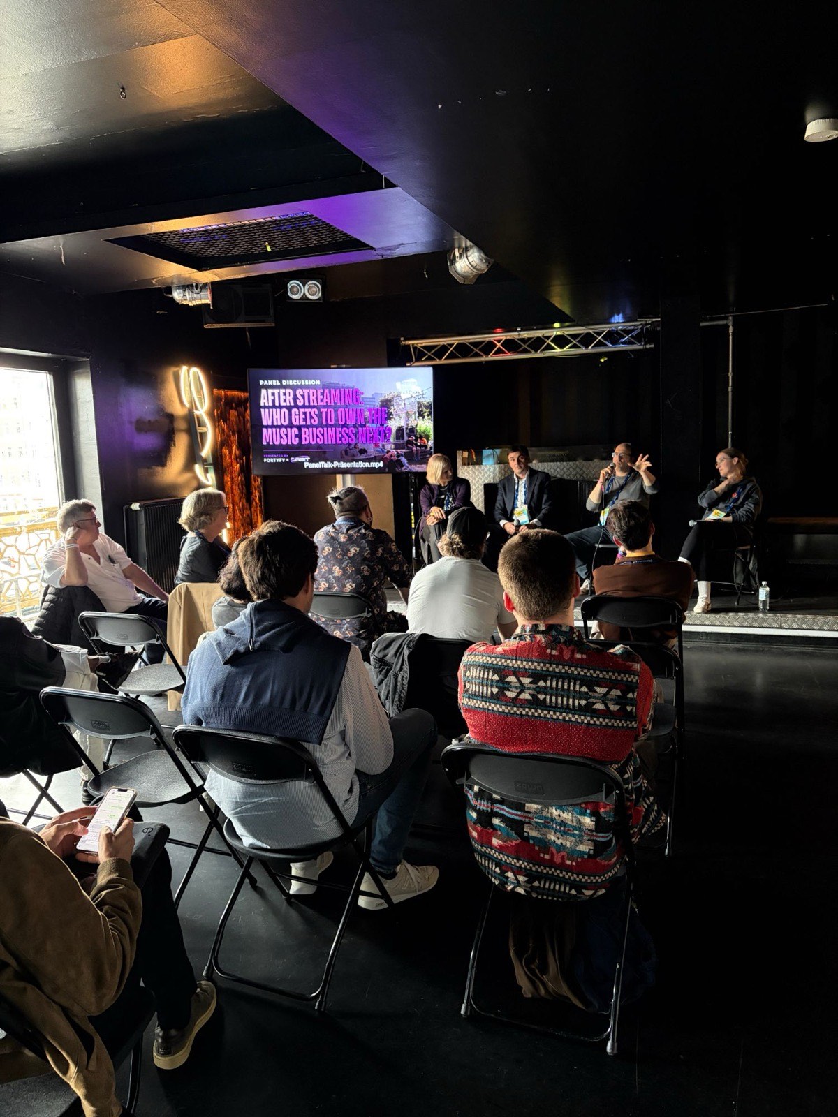 The panel seen from the floor at the side of the room, a speaker mid-gesture with the microphone and the audience in the foreground